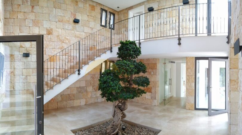 Imposantes Eingangsfoyer der Neubau Villa mit Natursteinwänden, geschwungener Treppe und dekorativem Bonsai-Baum.