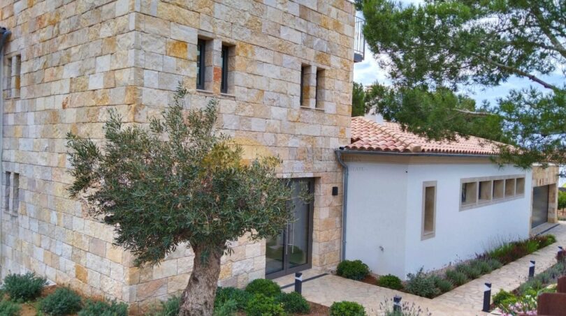 Außenansicht Neubau Villa Sol de Mallorca: Natursteinwände, weißer Anbau mit Terrakottadach, Olivenbaum und mediterraner Bepflanzung.