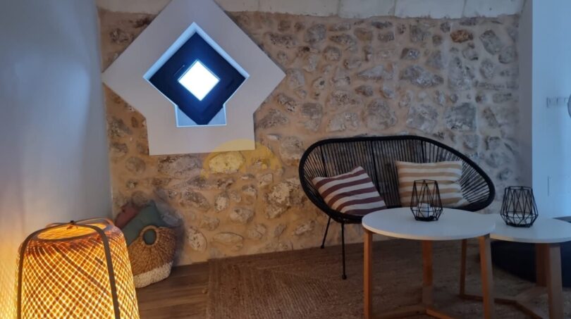 Gemütlicher Wohnbereich der Finca in Felanitx mit Steinwand, Rattanstuhl, Bambuslampe und rautenförmigem Fenster.