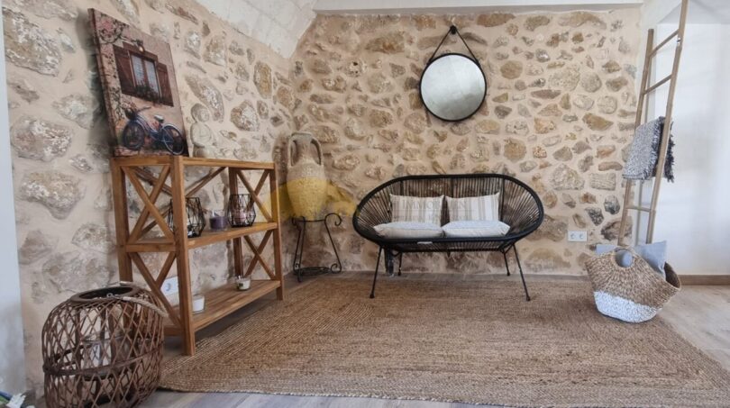 Rustikaler Wohnbereich der Finca Felanitx mit Natursteinwand, Rattansofa, Holzregal und dekorativen Elementen.