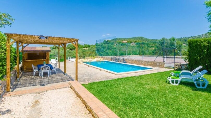 Außenbereich der Finca in Son Macià mit Pool, Pergola, Loungechairs, Tennisplatz und grünem Rasen.