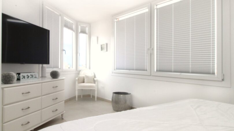 Helles Schlafzimmer des Strandpenthouses mit weißen Jalousien, TV-Kommode und elegantem Sessel