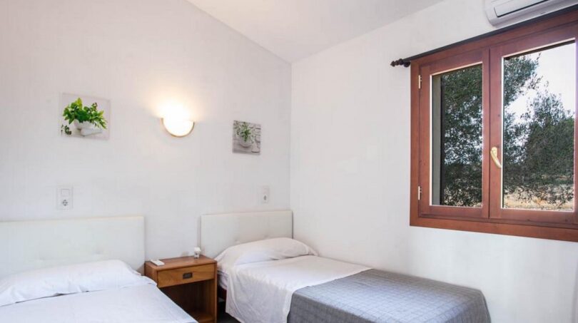Luminoso dormitorio de la finca Porto Cristo con dos camas individuales, aire acondicionado y ventanas de madera con vista a los árboles.