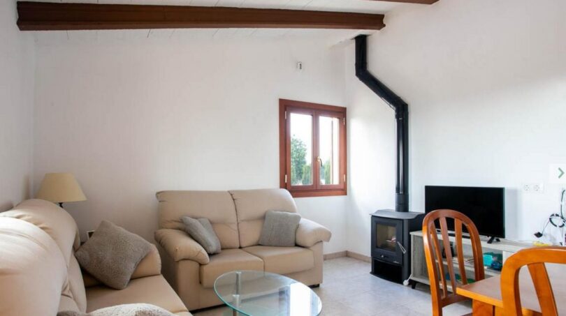Sala de estar de la finca en Porto Cristo con sofás, techo de vigas de madera, chimenea y ventanas con vistas a la naturaleza.