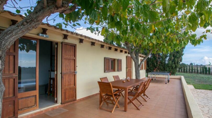 Soleada terraza de la finca Porto Cristo con muebles de madera, zona de barbacoa y árbol que proporciona sombra.