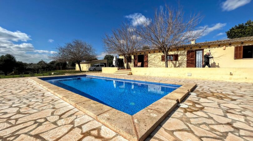 Finca Porto Cristo: Piscina rectangular con terraza de piedra natural y casa de campo mediterránea al fondo.