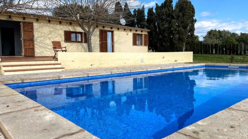 Finca Porto Cristo: Gran piscina exterior rectangular con agua azul, borde de piedra natural y casa de campo mediterránea al fondo.