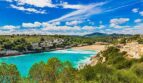 Bahía de agua turquesa de Porto Cristo con playa de arena, villas y paisaje de colinas verdes bajo cielo azul