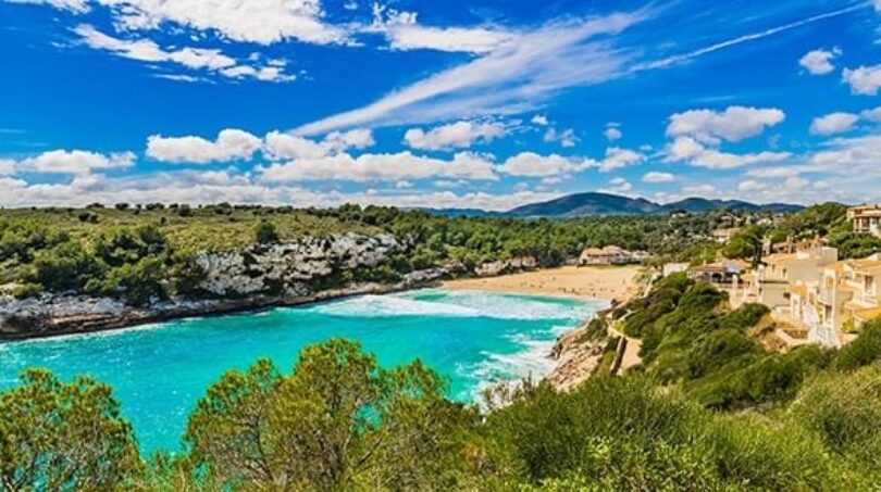 Bahía de agua turquesa de Porto Cristo con playa de arena, villas y paisaje de colinas verdes bajo cielo azul
