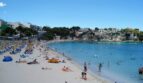 Concurrida playa de arena de Porto Cristo con agua turquesa, acantilados y pueblo al fondo.