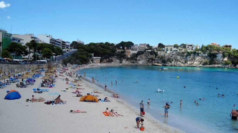 Concurrida playa de arena de Porto Cristo con agua turquesa, acantilados y pueblo al fondo.