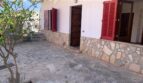 Außenansicht Zweifamilienhaus Portocolom: Terrasse mit Natursteinboden, weiße Fassade, rote Fensterläden und Holztür.