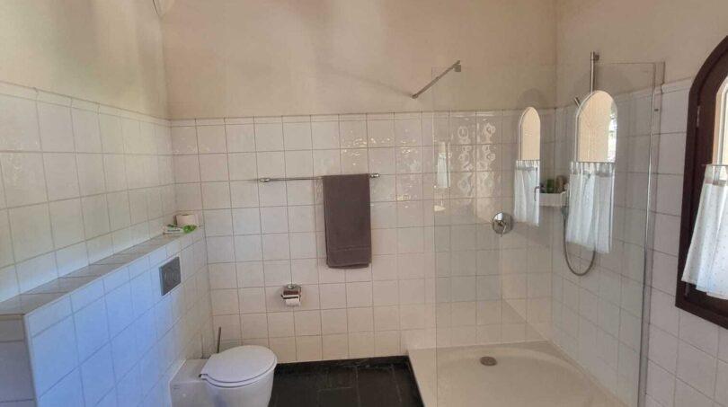 Baño luminoso de la villa con azulejos blancos, ducha, WC, bidé y vigas de madera rústicas en el techo.