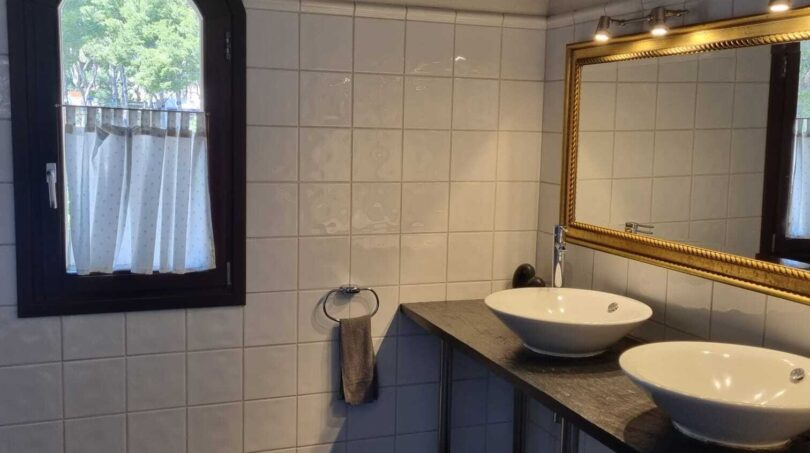 Elegante baño de la villa con dos lavabos sobre encimera, espejo dorado y ventana de arco redondo