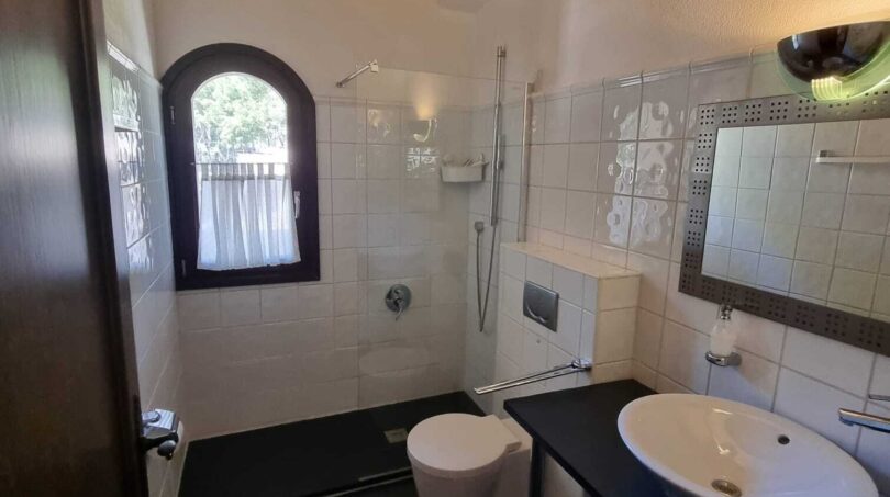Baño de la villa con ducha, WC, lavamanos, techo de vigas de madera y ventana de arco con vistas al jardín
