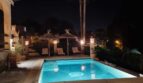 Piscina exterior iluminada de noche con tumbonas, techos de paja y palmeras en Villa Calvià