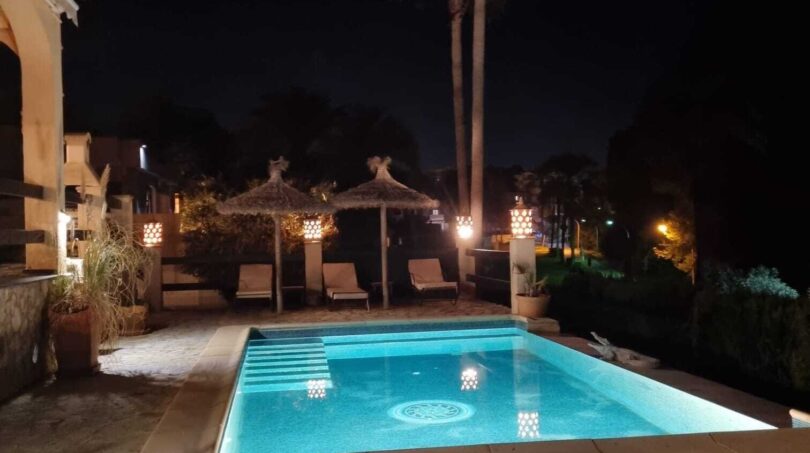 Piscina exterior iluminada de noche con tumbonas, techos de paja y palmeras en Villa Calvià