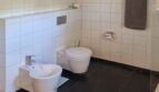 Baño moderno de la villa con WC mural, bidé, suelo de mármol negro y azulejos blancos en la pared.