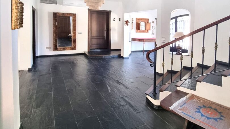 Zona de entrada de la villa con suelo de piedra natural oscura, escalera de madera, araña y elegante portal de madera.