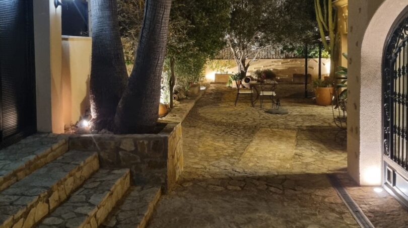 Patio interior iluminado de una villa exclusiva en Calvia por la noche con adoquines de piedra natural, palmeras y olivo.