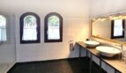 Espacioso baño de la villa con dos lavabos, techo de vigas de madera, suelo de piedra natural y ventanas de arco