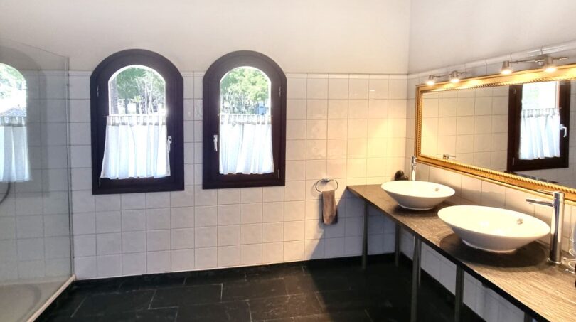 Espacioso baño de la villa con dos lavabos, techo de vigas de madera, suelo de piedra natural y ventanas de arco
