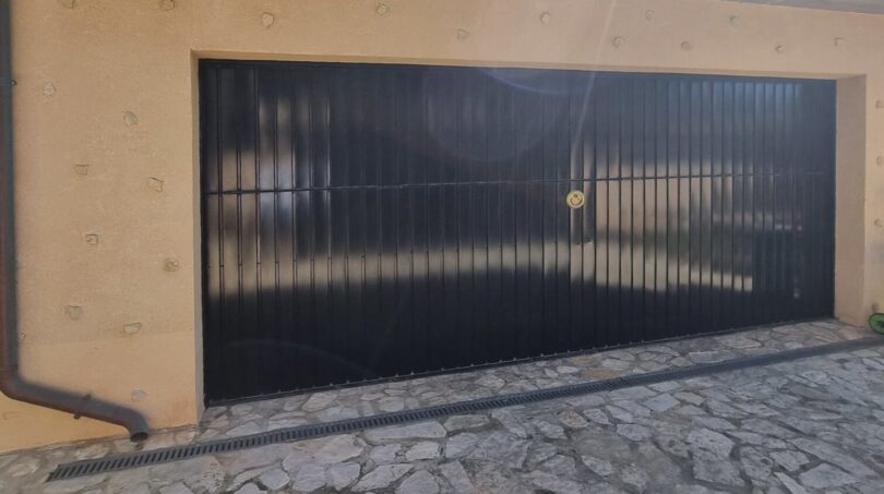 Garaje doble con puerta de lamas negra, pavimento de piedra natural y techo de tejas de terracota mediterránea de la villa en Costa de la Calma