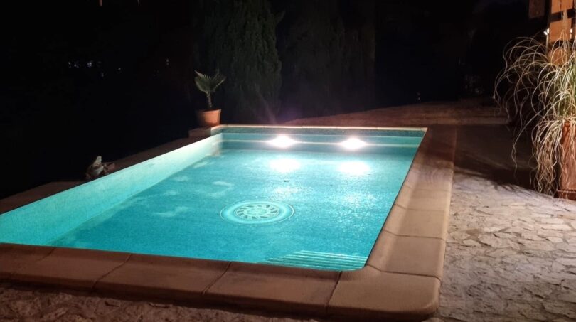 Piscina iluminada de noche con borde de piedra natural y palapa, villa en Costa de la Calma