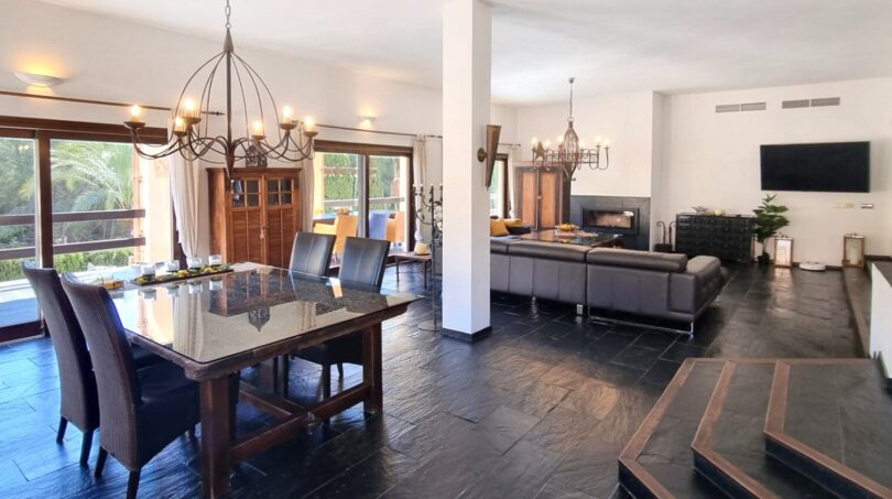 Zona de estar y comedor abierta con suelo de pizarra oscura, mesa de cristal y elegante araña en villa exclusiva
