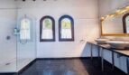 Baño luminoso de la villa en Calvià con ducha de cristal, dos lavabos sobre encimera, ventanas de arco y suelo de piedra oscura.