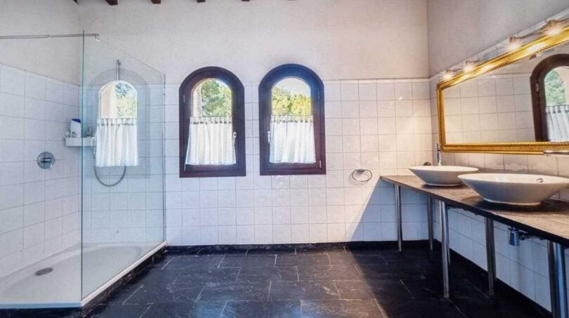 Baño luminoso de la villa en Calvià con ducha de cristal, dos lavabos sobre encimera, ventanas de arco y suelo de piedra oscura.