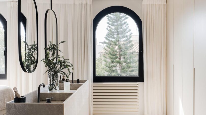 Elegantes Badezimmer der Maisonette mit Designspiegel, Naturstein-Waschtisch und Bogenfenster mit Meerblick