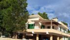 Zweistöckige Villa in Alcúdia mit Balkon, Carport, weißer Fassade und großem Kiefernbaum vor blauem Himmel.