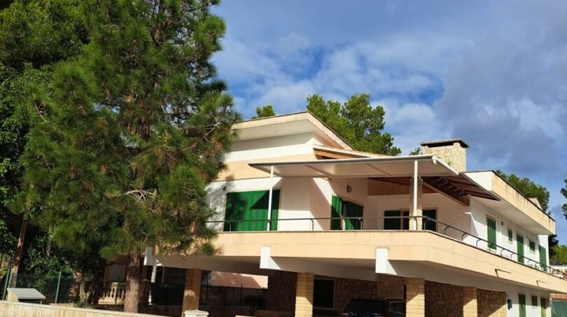 Zweistöckige Villa in Alcúdia mit Balkon, Carport, weißer Fassade und großem Kiefernbaum vor blauem Himmel.
