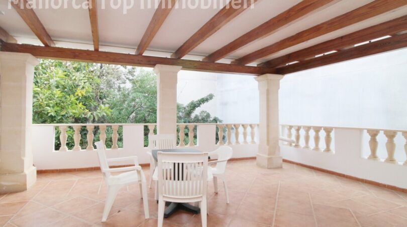 Überdachte Terrasse der Villa in Alcúdia mit Holzbalken, Säulen, weißen Gartenmöbeln und Gartenblick.