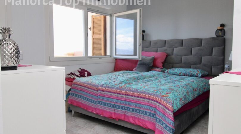 Helles Schlafzimmer mit Doppelbett, grauem Polsterkopfteil und bunter Bettwäsche, Fenster mit Meerblick