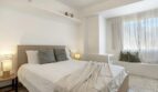 Helles Schlafzimmer mit Doppelbett, grau-weißen Kissen, Wandregal und Sitzbank am Fenster in Palma-Wohnung.