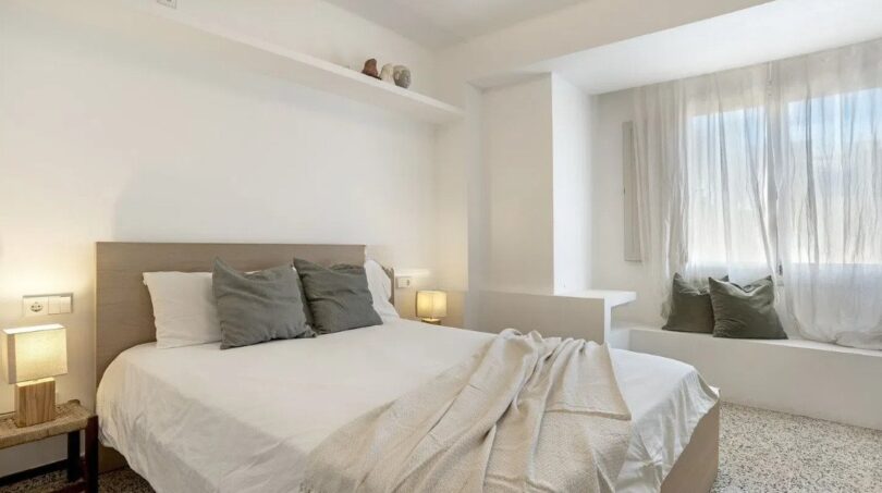 Helles Schlafzimmer mit Doppelbett, grau-weißen Kissen, Wandregal und Sitzbank am Fenster in Palma-Wohnung.