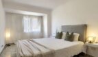 Helles Schlafzimmer mit Doppelbett, grauem Polsterkopfteil und weißen Wänden in moderner Wohnung in Palma de Mallorca.
