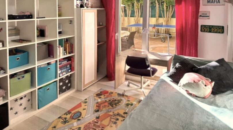 Kinderzimmer mit Regalwand, Schreibtisch, Spielteppich und Zugang zur sonnigen Terrasse mit Bäumen