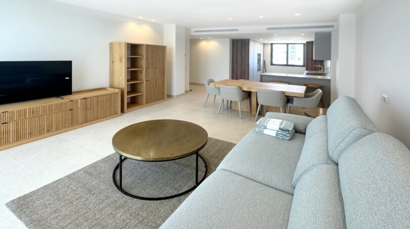 Helles Wohnzimmer mit grauem Sofa, rundem Couchtisch und offener Küche in exklusiver Erstbezugswohnung in Palma