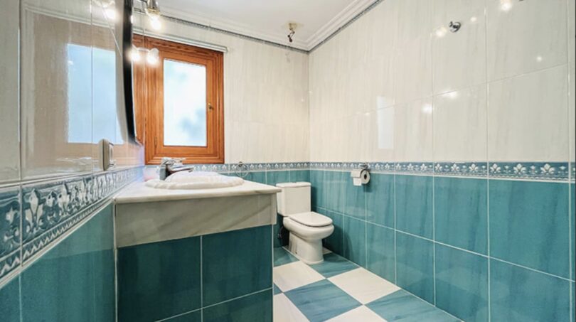 Helles Badezimmer der Villa in Cala Vinyes mit blauen Fliesen, Waschbecken, WC und Holzfenster.