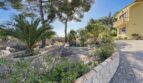 Gepflegter Villengarten in Cala Vinyes mit Palmen, Steinbeet und mediteraner Bepflanzung neben gelbem Villengebäude.