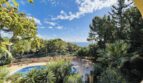 Außenbereich der Villa in Cala Vinyes: Runder Pool, üppiger Garten mit Palmen und Meerblick bei blauem Himmel.