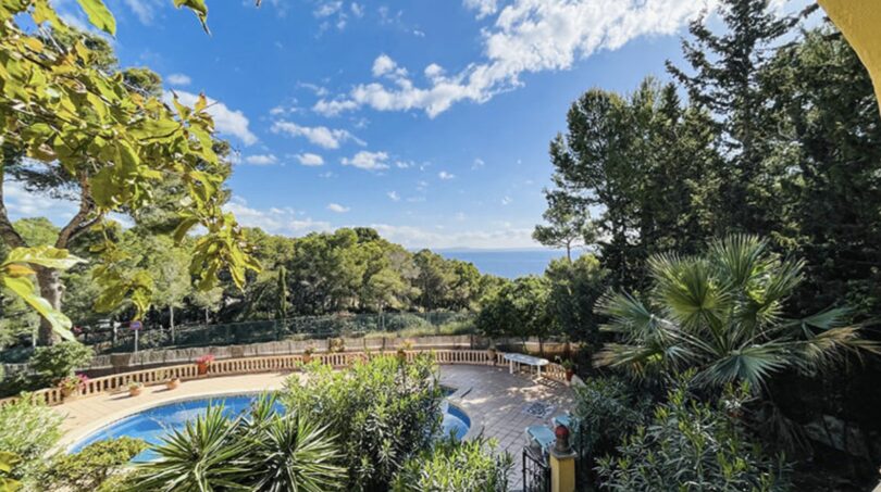 Außenbereich der Villa in Cala Vinyes: Runder Pool, üppiger Garten mit Palmen und Meerblick bei blauem Himmel.