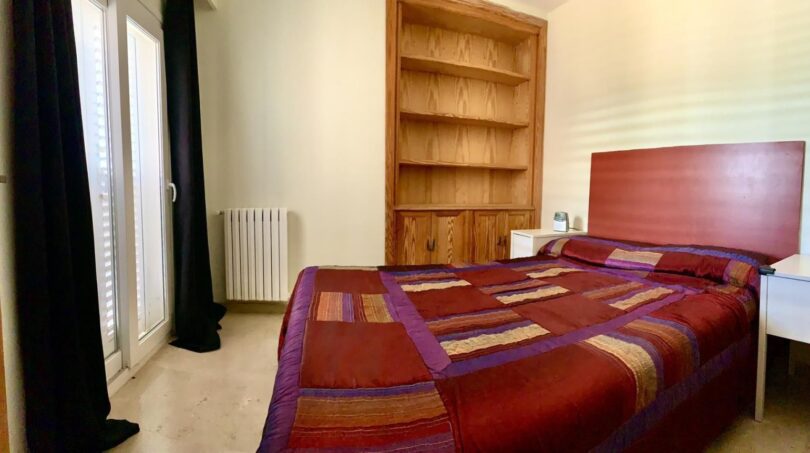 Schlafzimmer des Chalets in Marratxí mit Doppelbett, Holzregal, Heizkörper und Balkontür mit dunklen Vorhängen.