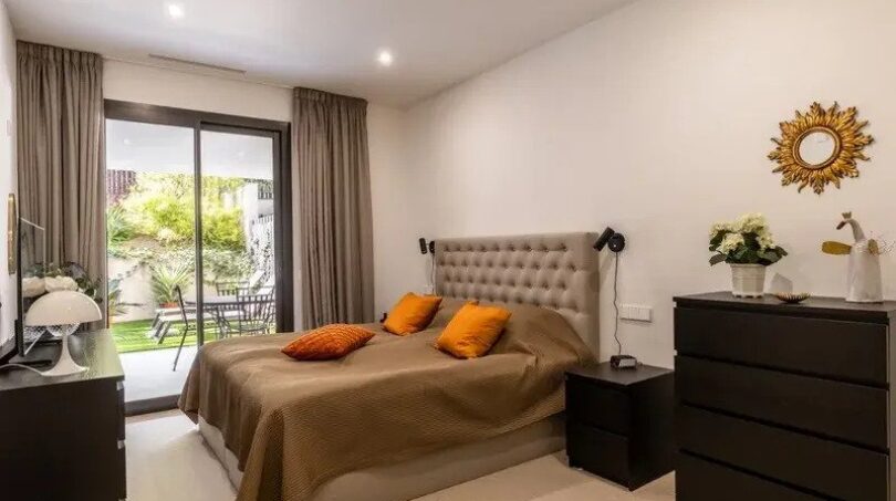Bright bedroom of the ground-floor apartment La Vileta with double bed, orange cushions and terrace access to the garden.
