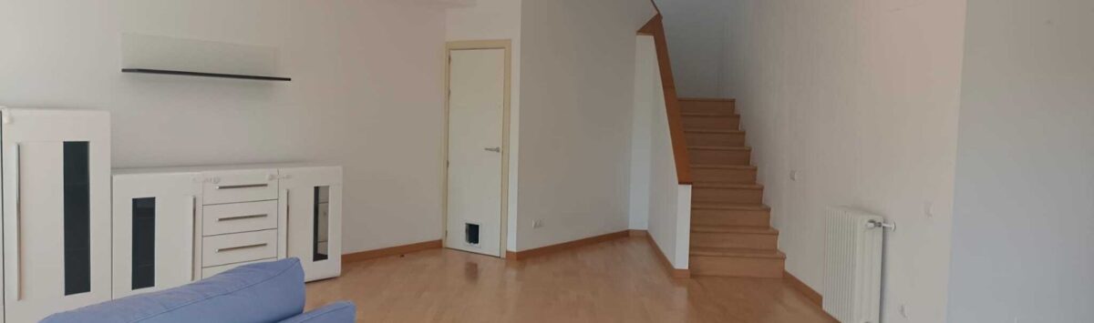 Salón luminoso con suelo de madera, sofá azul, cómoda blanca y escalera abierta a la planta superior.