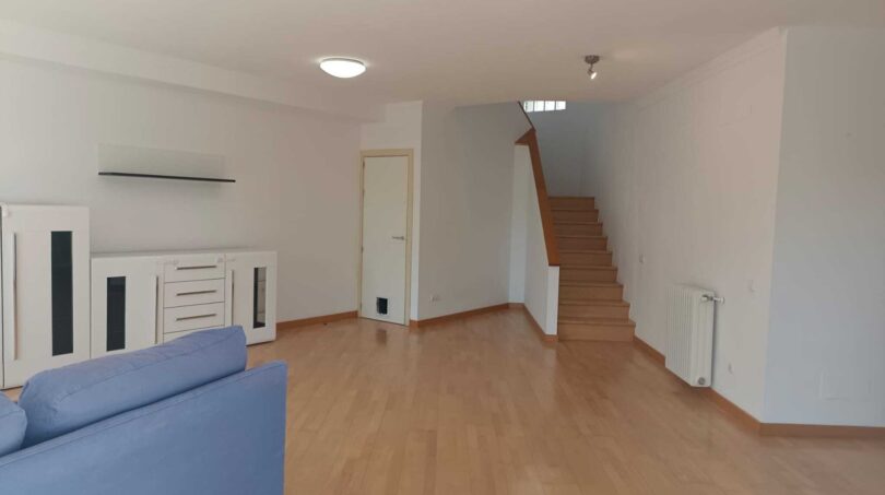 Bright living room with wooden floor, blue sofa, white dresser and open staircase to the upper floor.