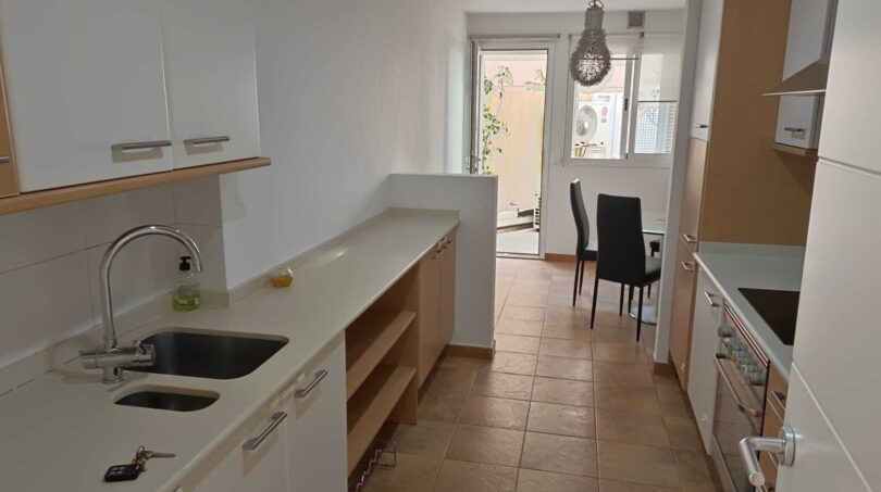 Galley kitchen with white high-gloss fronts, double sink and view of dining area with terrace access.